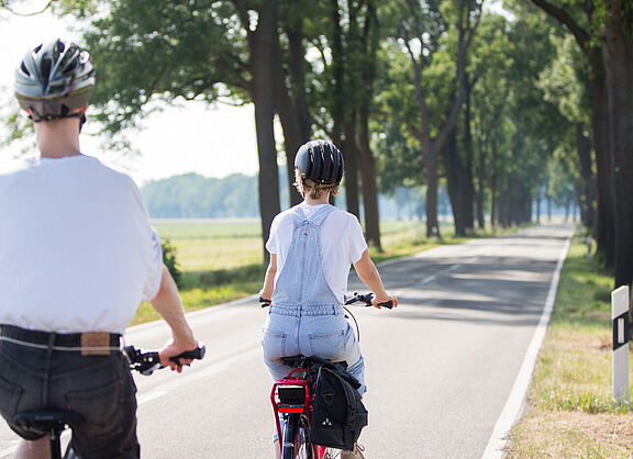 Zwei Personen mit Fahrradhelmen fahren auf einer schmalen Landstraße ohne separaten Radweg.